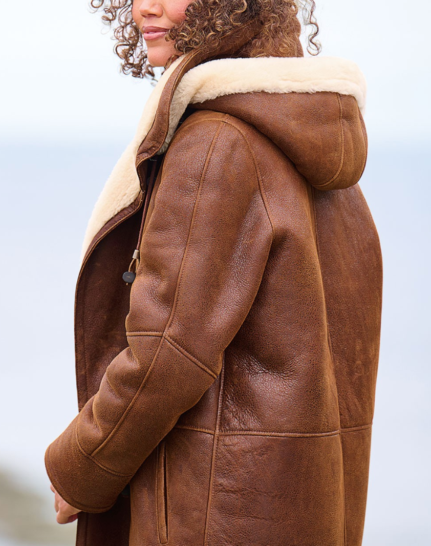 Brown - A woman wearing a mid-length brown textured suede-look coat with a plush cream shearling lining, featuring a hood, toggle closures, and a front zipper.