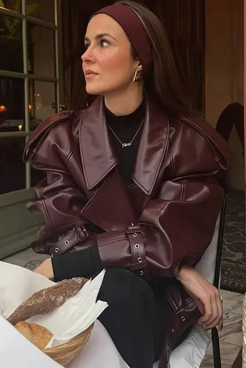 Woman wearing an oversized burgundy faux leather moto jacket with exaggerated shoulders, wide collar, and belted cuffs, styled in a chic city setting.