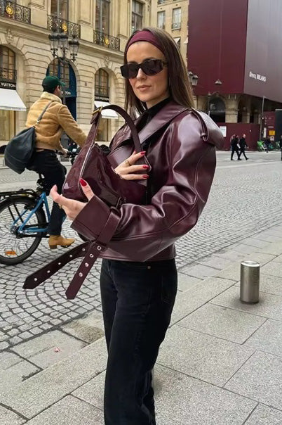 Woman wearing an oversized burgundy faux leather moto jacket with exaggerated shoulders, wide collar, and belted cuffs, styled in a chic city setting.