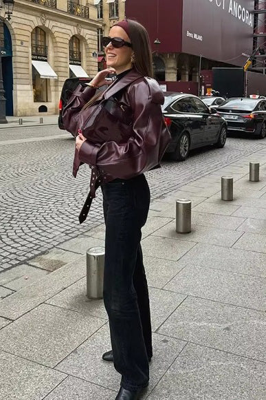 Woman wearing an oversized burgundy faux leather moto jacket with exaggerated shoulders, wide collar, and belted cuffs, styled in a chic city setting.