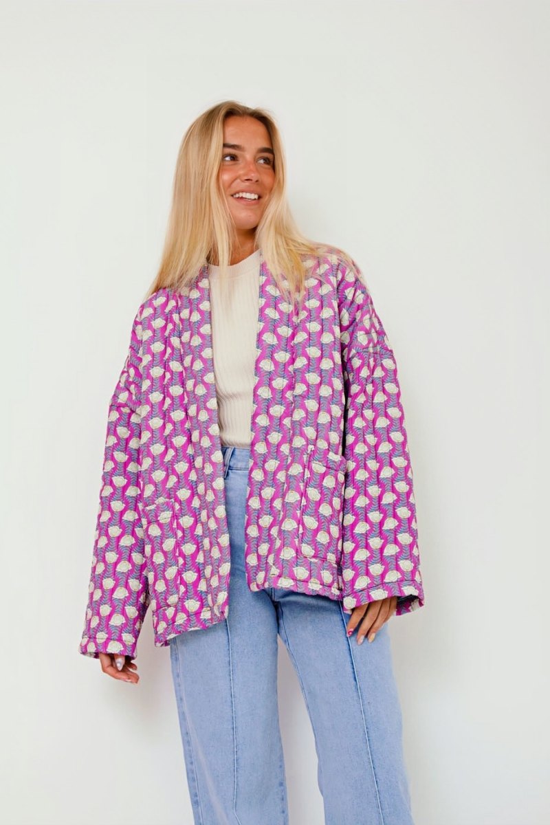 Woman wearing a pink quilted mid-length jacket with a white floral pattern, styled open over a white top and light blue jeans.