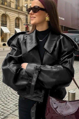 Woman wearing an oversized burgundy faux leather moto jacket with exaggerated shoulders, wide collar, and belted cuffs, styled in a chic city setting.