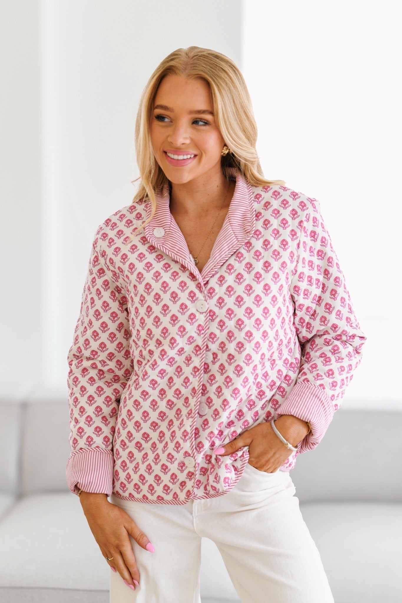 Pink - A woman wearing a white quilted jacket with a pink floral pattern and pink striped collar and cuffs, paired with white jeans.