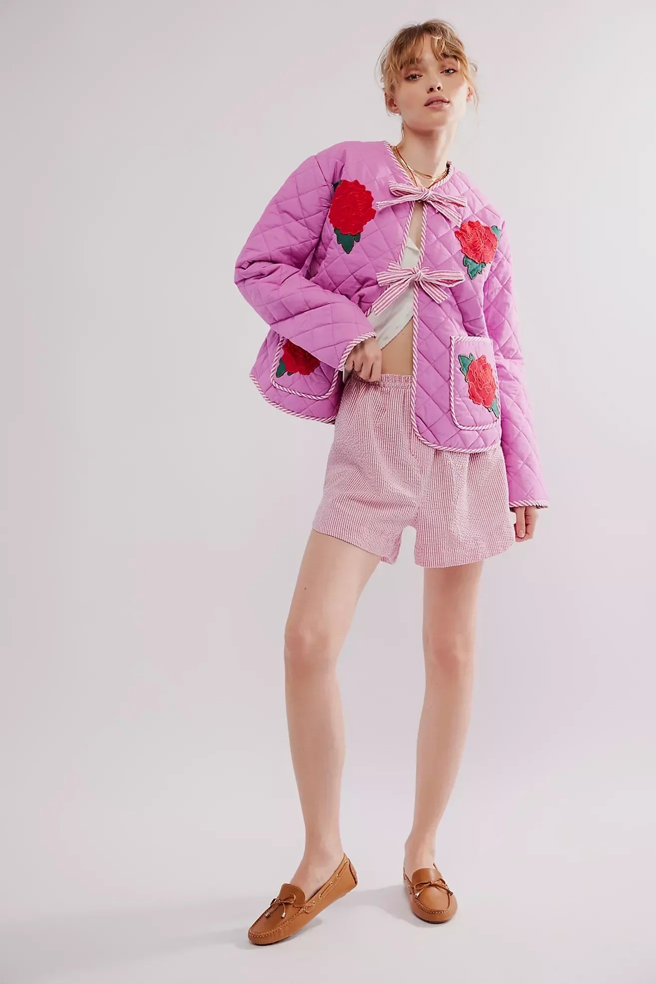 Women's pink diamond-quilted jacket featuring large red rose appliqués, striped tie-front closures, and matching piped edges, worn over a white top and pink striped shorts.