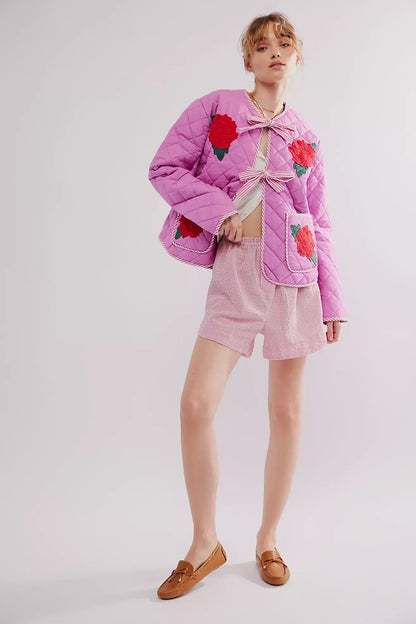 Women's pink diamond-quilted jacket featuring large red rose appliqués, striped tie-front closures, and matching piped edges, worn over a white top and pink striped shorts.