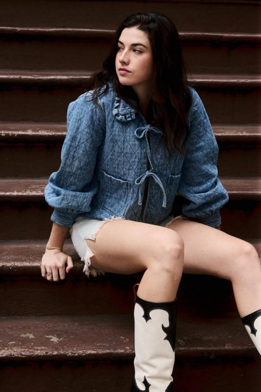 Women's blue diamond-quilted denim jacket featuring a ruffled collar, tie-front closure, and voluminous balloon sleeves, styled with distressed shorts and cowboy boots.