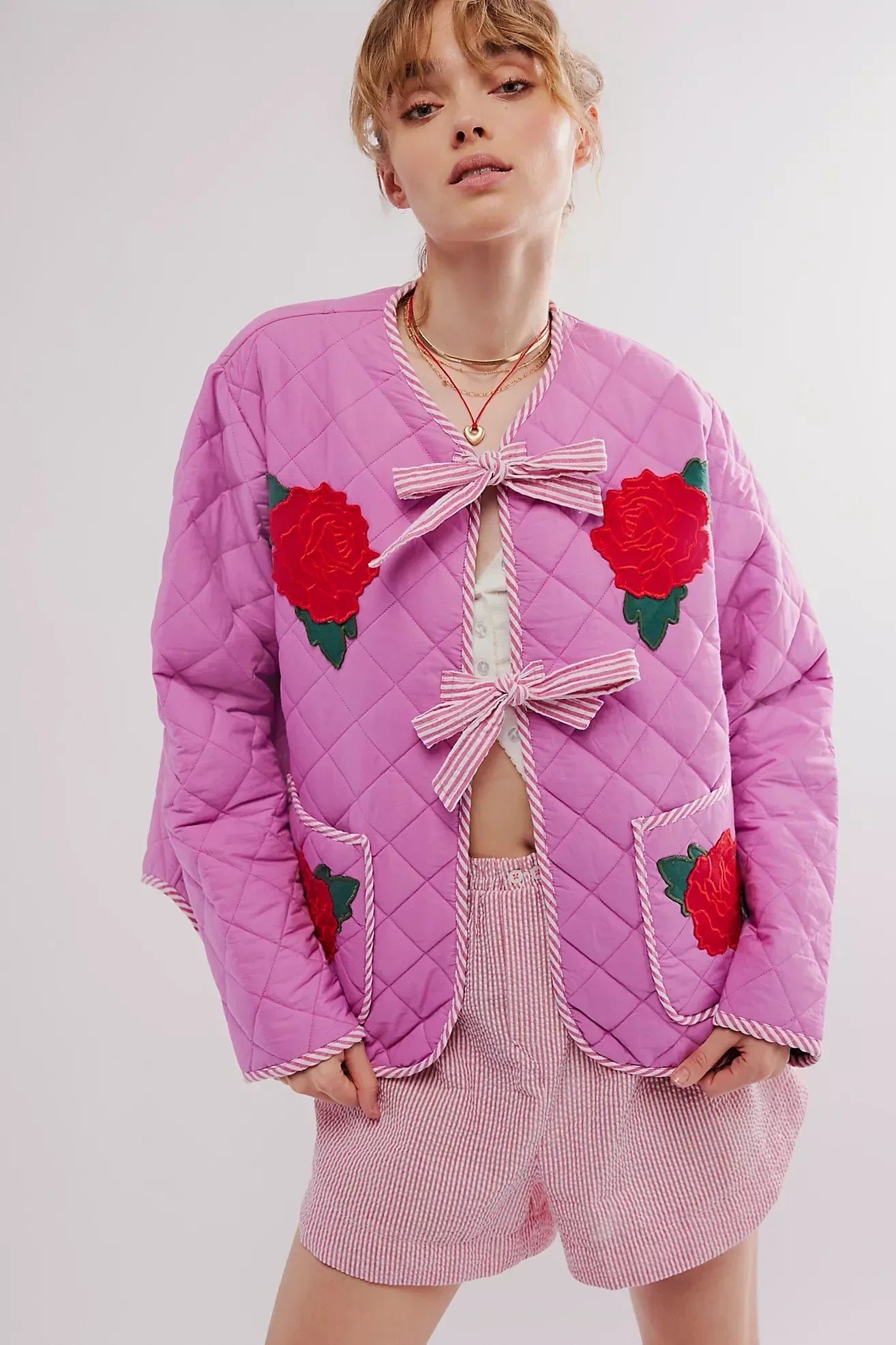 Women's pink diamond-quilted jacket featuring large red rose appliqués, striped tie-front closures, and matching piped edges, worn over a white top and pink striped shorts.