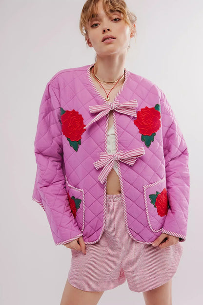 Women's pink diamond-quilted jacket featuring large red rose appliqués, striped tie-front closures, and matching piped edges, worn over a white top and pink striped shorts.
