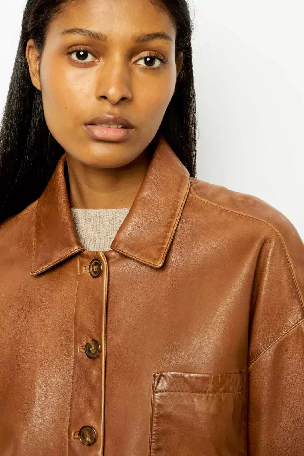 Brown - A woman wearing a relaxed-fit, brown faux leather button-front jacket with a classic collar and single chest pocket, styled over a patterned knit sweater.