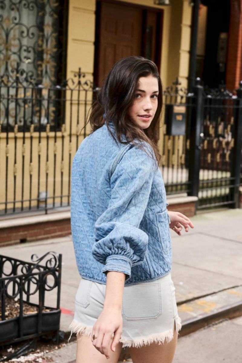 Women's blue diamond-quilted denim jacket featuring a ruffled collar, tie-front closure, and voluminous balloon sleeves, styled with distressed shorts and cowboy boots.