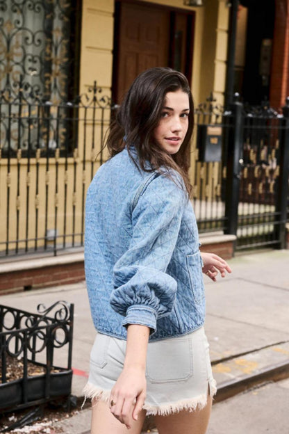 Women's blue diamond-quilted denim jacket featuring a ruffled collar, tie-front closure, and voluminous balloon sleeves, styled with distressed shorts and cowboy boots.
