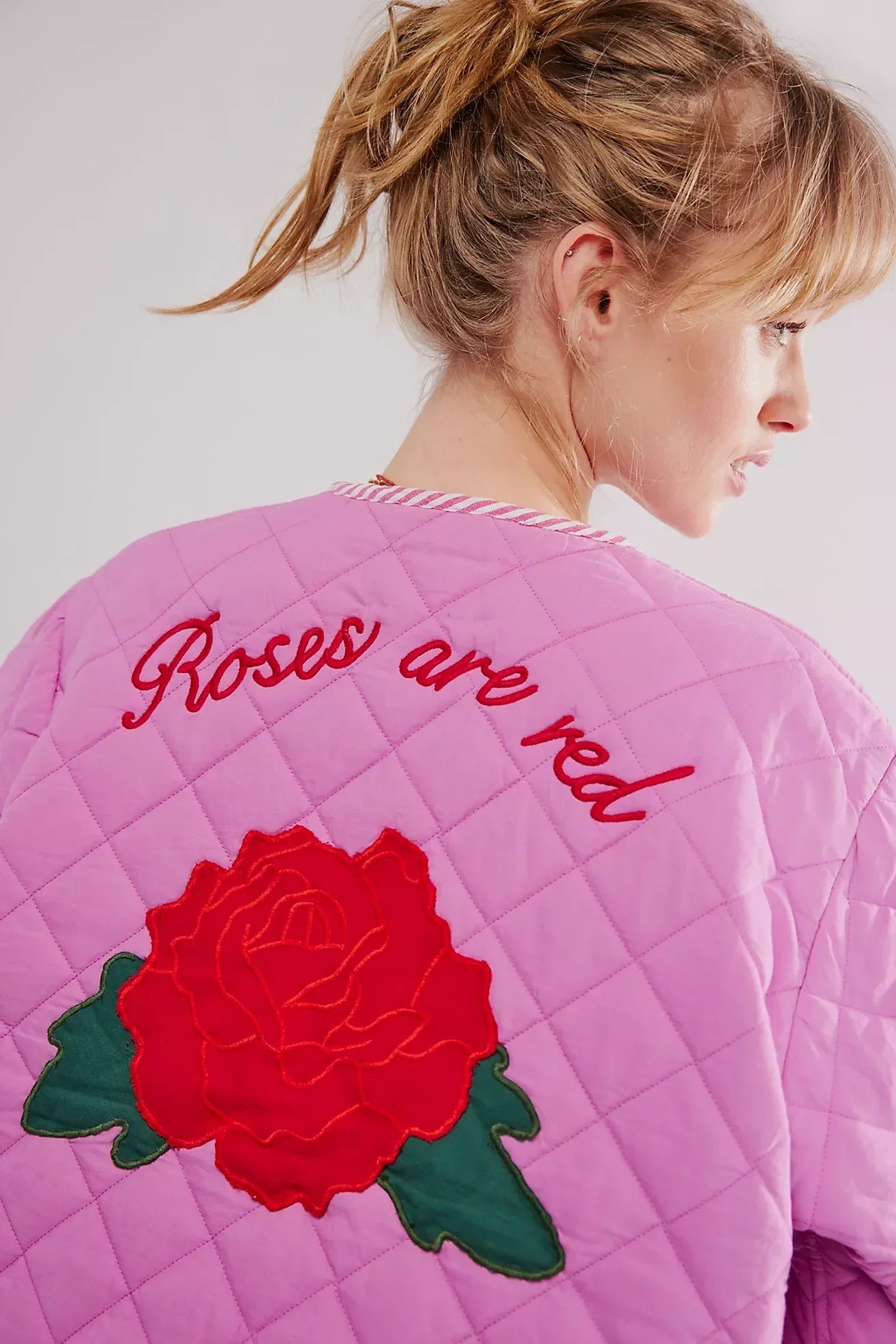 Women's pink diamond-quilted jacket featuring large red rose appliqués, striped tie-front closures, and matching piped edges, worn over a white top and pink striped shorts.