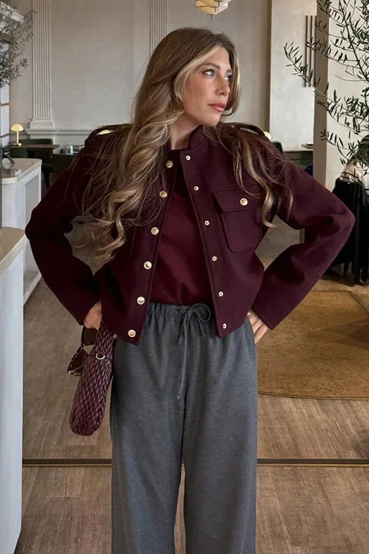 Wine Red - A woman wearing a deep burgundy, military-inspired cropped jacket with gold-tone buttons and chest pockets, paired with grey trousers.