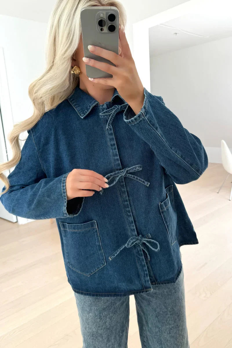 A woman taking a mirror selfie wearing a blue denim jacket featuring three tie-front closures and patch pockets, paired with wide-leg denim trousers.