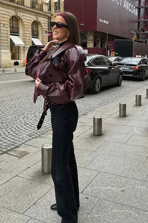 Woman wearing an oversized burgundy faux leather moto jacket with exaggerated shoulders, wide collar, and belted cuffs, styled in a chic city setting.