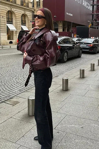 Woman wearing an oversized burgundy faux leather moto jacket with exaggerated shoulders, wide collar, and belted cuffs, styled in a chic city setting.