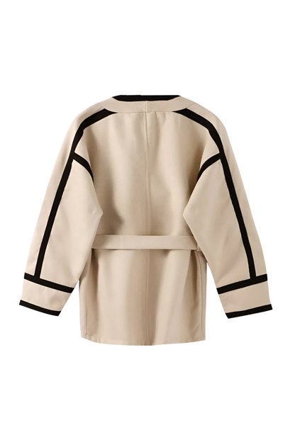 Khaki - A woman wearing a tan collarless wrap jacket with bold black contrast trim and a self-tie belt, layered over a blue striped shirt and jeans.