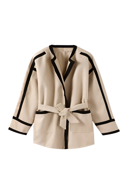 Khaki - A woman wearing a tan collarless wrap jacket with bold black contrast trim and a self-tie belt, layered over a blue striped shirt and jeans.
