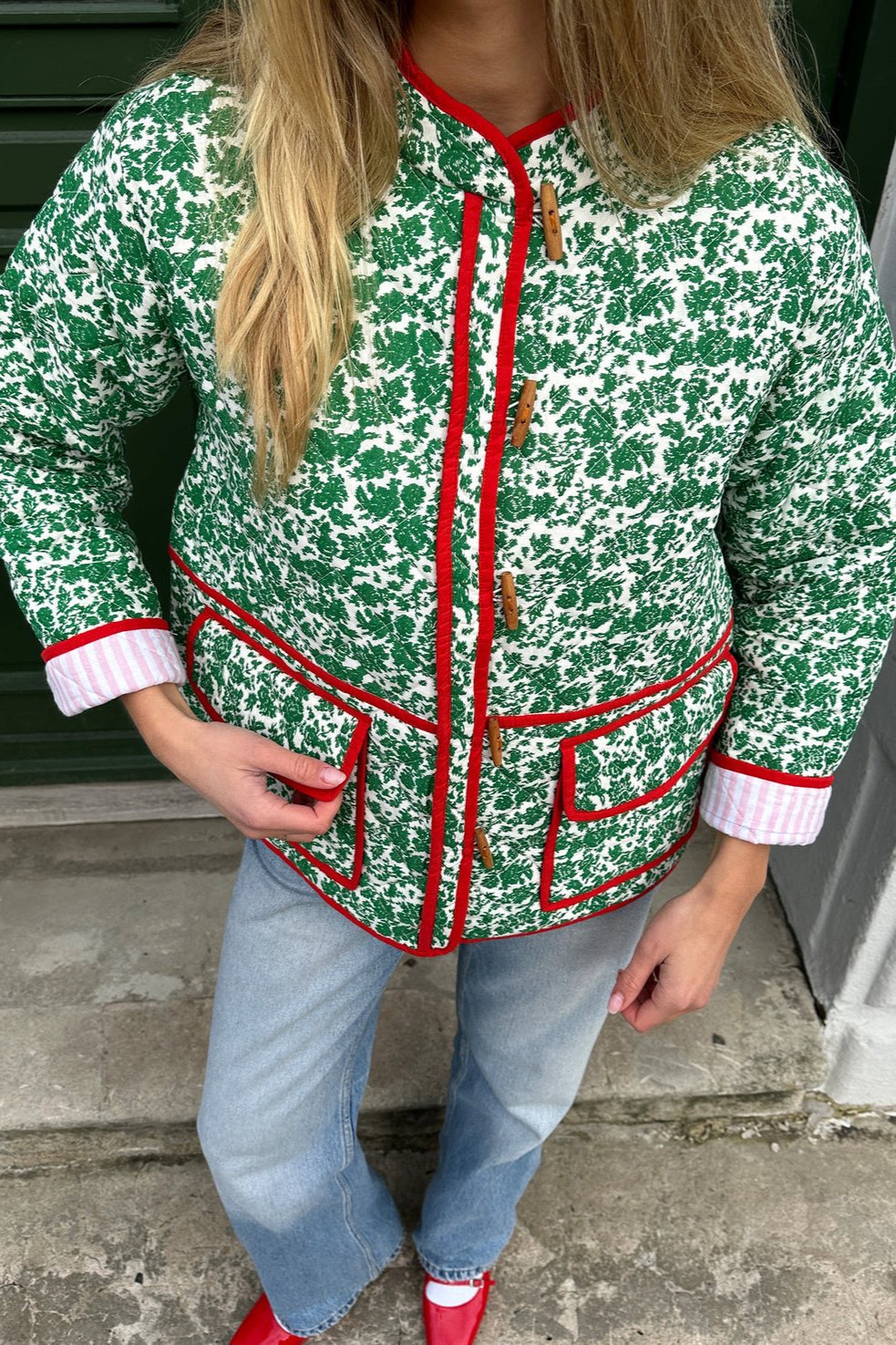 Woman wearing a green and white ditsy floral quilted jacket with red piped trim over a pink striped dress.