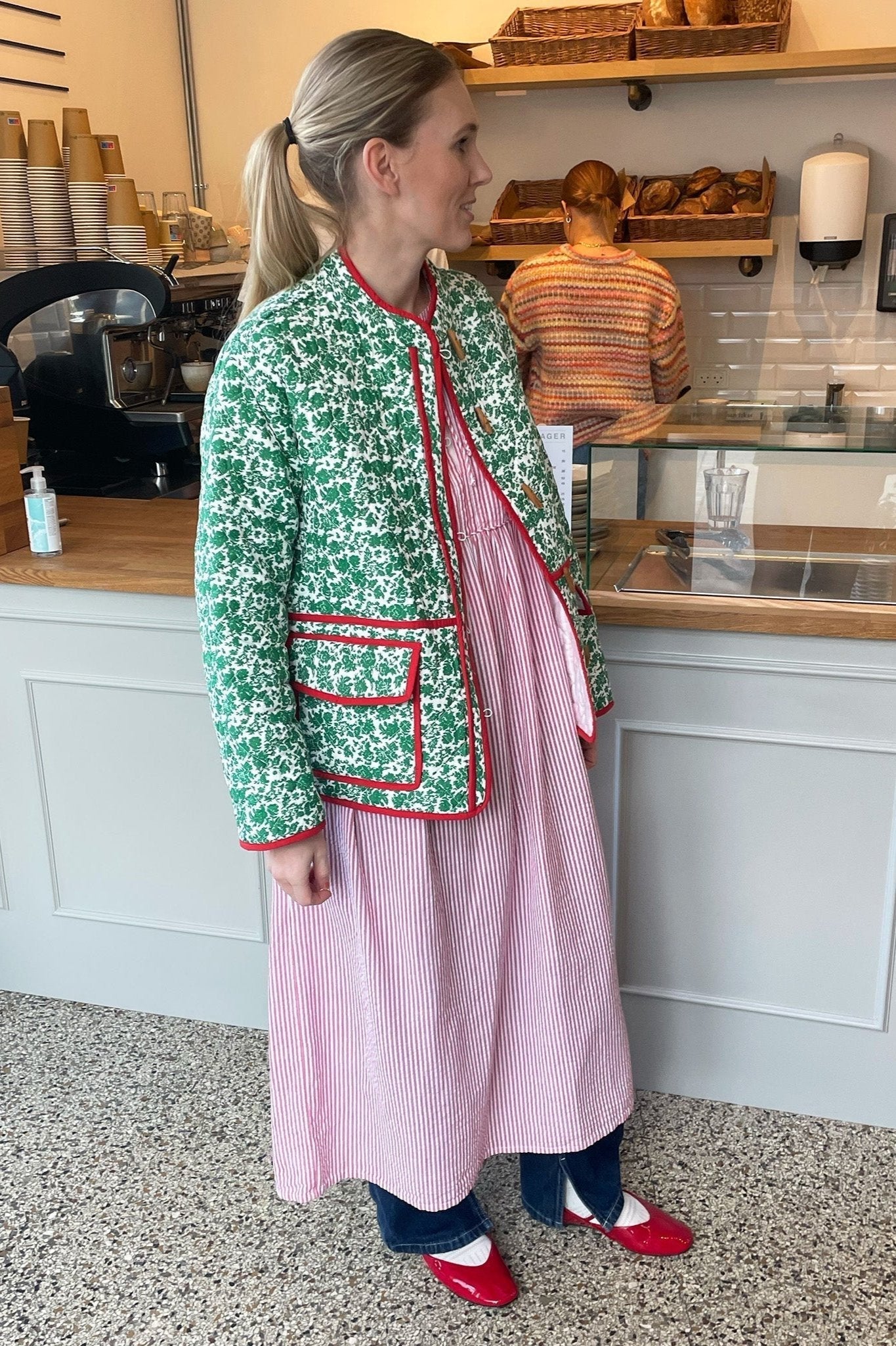 Woman wearing a green and white ditsy floral quilted jacket with red piped trim over a pink striped dress.