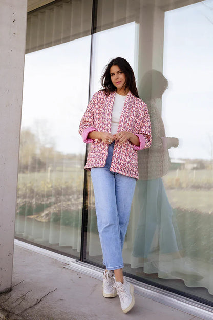 Woman wearing a pink and red geometric print quilted open-front jacket with rolled cuffs, styled with light wash jeans and a white tee.
