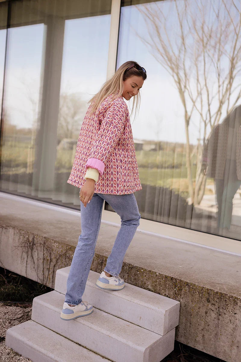 Woman wearing a pink and red geometric print quilted open-front jacket with rolled cuffs, styled with light wash jeans and a white tee.