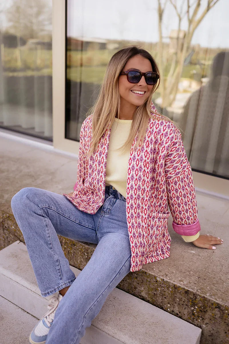Woman wearing a pink and red geometric print quilted open-front jacket with rolled cuffs, styled with light wash jeans and a white tee.