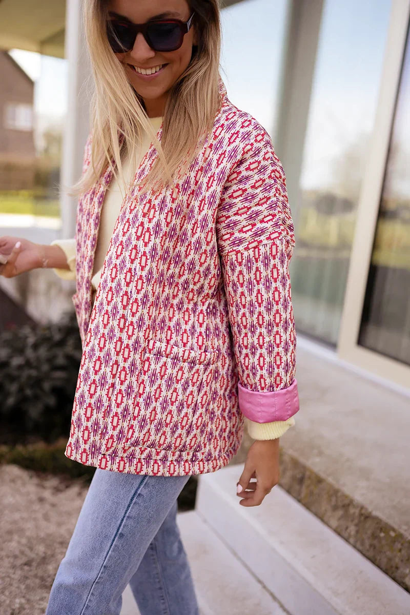 Woman wearing a pink and red geometric print quilted open-front jacket with rolled cuffs, styled with light wash jeans and a white tee.