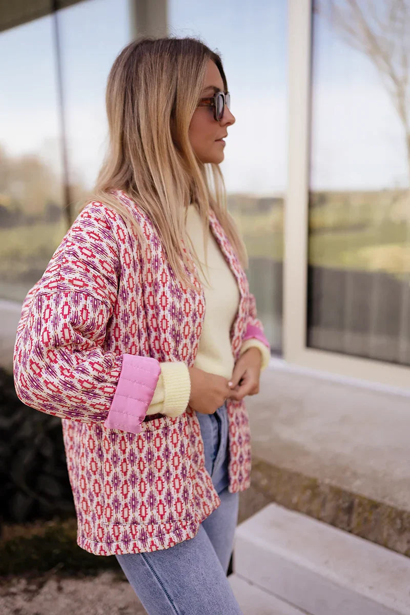Woman wearing a pink and red geometric print quilted open-front jacket with rolled cuffs, styled with light wash jeans and a white tee.