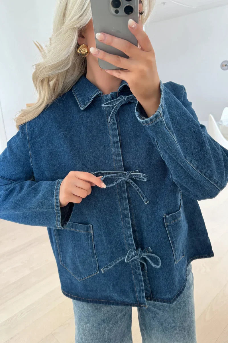 A woman taking a mirror selfie wearing a blue denim jacket featuring three tie-front closures and patch pockets, paired with wide-leg denim trousers.