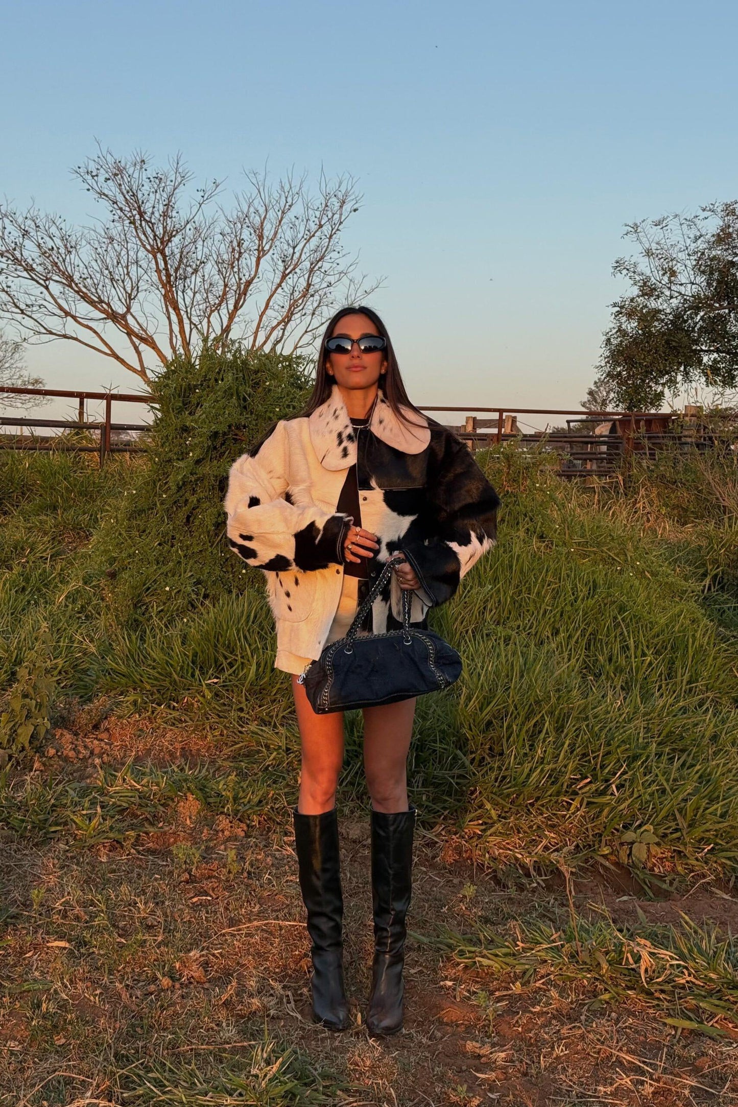 Woman standing outdoors wearing an oversized black and white cow print faux fur jacket, styled with a plaid shirt tied around her waist and tall black boots.
