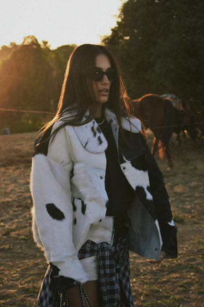Woman standing outdoors wearing an oversized black and white cow print faux fur jacket, styled with a plaid shirt tied around her waist and tall black boots.