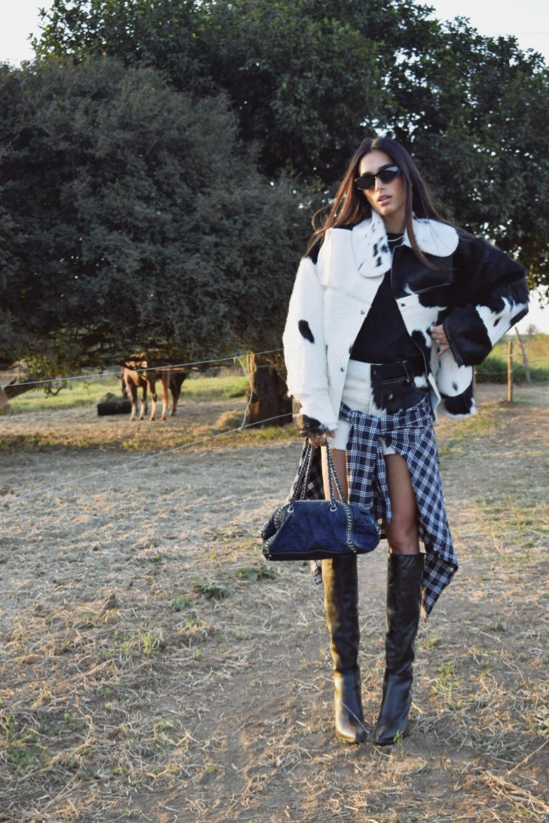 Milk - Spot White - Woman standing outdoors wearing an oversized black and white cow print faux fur jacket, styled with a plaid shirt tied around her waist and tall black boots.