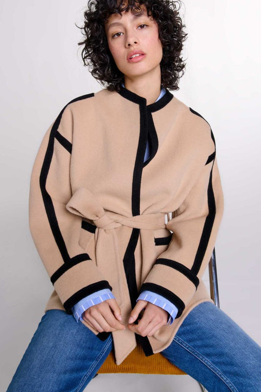 A woman wearing a tan collarless wrap jacket with bold black contrast trim and a self-tie belt, layered over a blue striped shirt and jeans.