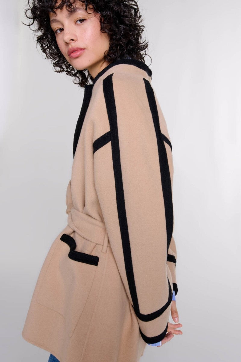 A woman wearing a tan collarless wrap jacket with bold black contrast trim and a self-tie belt, layered over a blue striped shirt and jeans.
