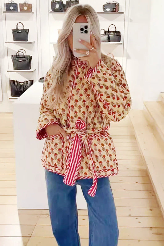 Woman taking a mirror selfie wearing a quilted, cream-coloured wrap jacket with a red floral block print and striped tie belt, paired with blue jeans.