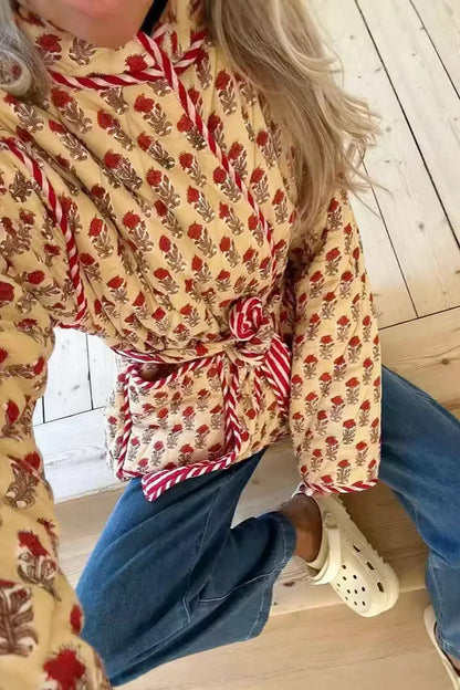 Woman taking a mirror selfie wearing a quilted, cream-coloured wrap jacket with a red floral block print and striped tie belt, paired with blue jeans.