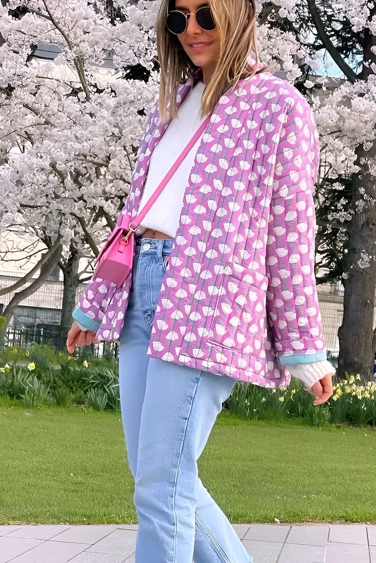 Woman wearing a pink quilted mid-length jacket with a white floral pattern, styled open over a white top and light blue jeans.