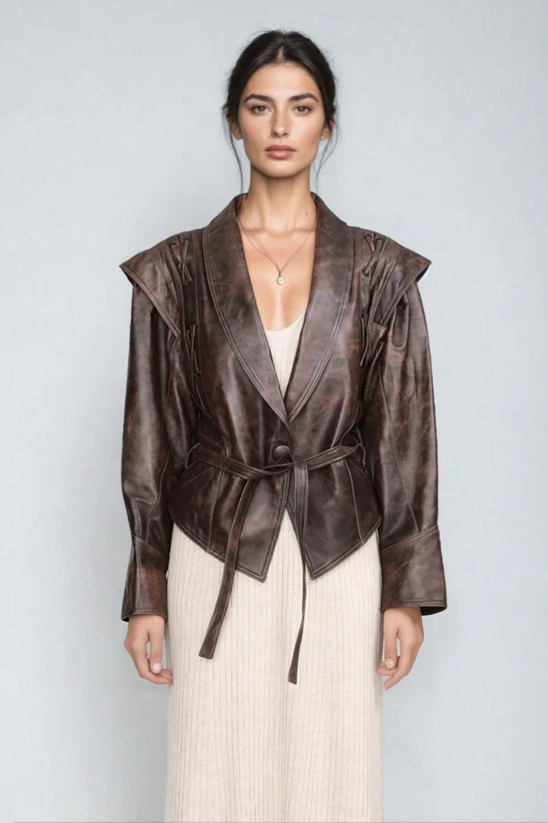 Brown - A woman wearing a brown distressed faux leather jacket with oversized shoulders, lacing details, and a tie belt at the waist, styled over a light ribbed dress.