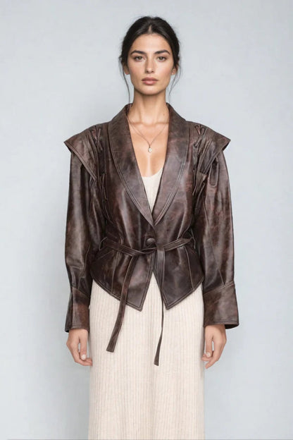 Brown - A woman wearing a brown distressed faux leather jacket with oversized shoulders, lacing details, and a tie belt at the waist, styled over a light ribbed dress.