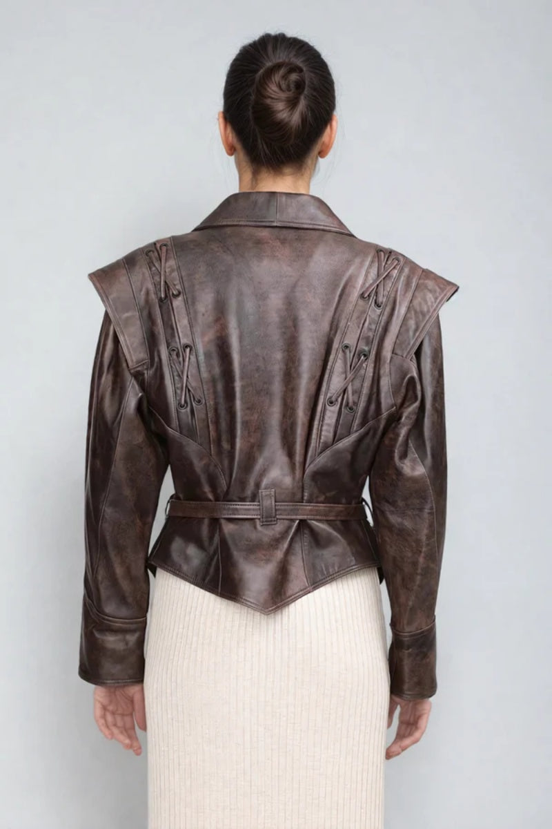 Brown - A woman wearing a brown distressed faux leather jacket with oversized shoulders, lacing details, and a tie belt at the waist, styled over a light ribbed dress.