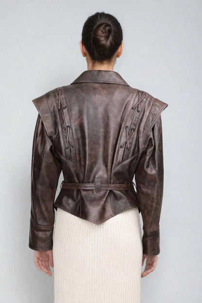 Brown - A woman wearing a brown distressed faux leather jacket with oversized shoulders, lacing details, and a tie belt at the waist, styled over a light ribbed dress.