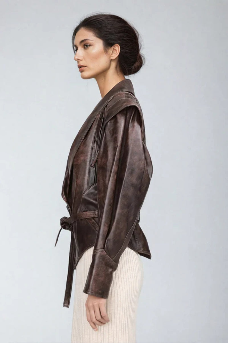 Brown - A woman wearing a brown distressed faux leather jacket with oversized shoulders, lacing details, and a tie belt at the waist, styled over a light ribbed dress.