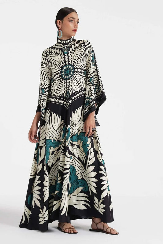 Multi - A woman wearing a high-neck maxi dress with a bold engineered botanical print in black, cream, and teal, featuring wide cape sleeves and a fluid, floor-length drape.