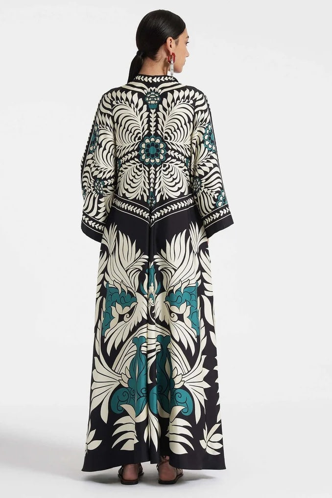 Multi - A woman wearing a high-neck maxi dress with a bold engineered botanical print in black, cream, and teal, featuring wide cape sleeves and a fluid, floor-length drape.