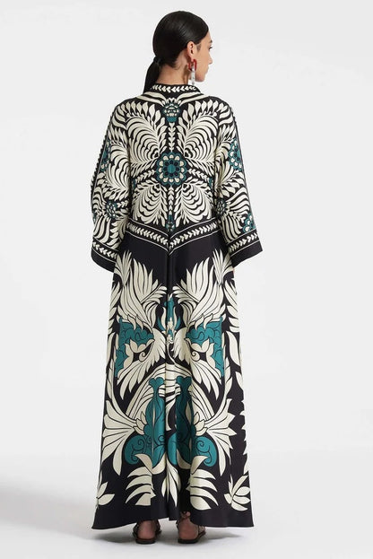 Multi - A woman wearing a high-neck maxi dress with a bold engineered botanical print in black, cream, and teal, featuring wide cape sleeves and a fluid, floor-length drape.