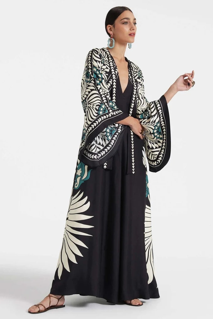 Multi - A woman wearing a high-neck maxi dress with a bold engineered botanical print in black, cream, and teal, featuring wide cape sleeves and a fluid, floor-length drape.