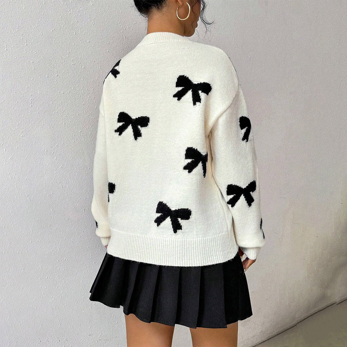 White - Women's relaxed fit white knit sweater featuring a scattered black bow pattern, styled with a black pleated mini skirt.