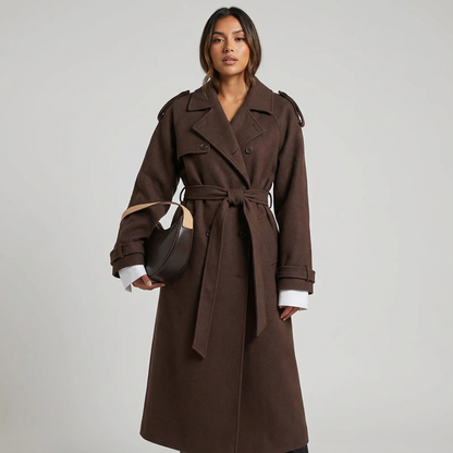 A woman wears a chocolate brown, double-breasted midi trench coat with a self-tie belt, styled over a white knit top and dark pants, holding a brown leather handbag.