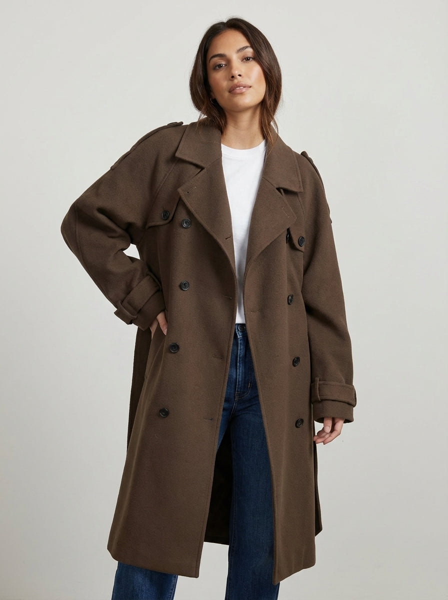 A woman wears a chocolate brown, double-breasted midi trench coat with a self-tie belt, styled over a white knit top and dark pants, holding a brown leather handbag.